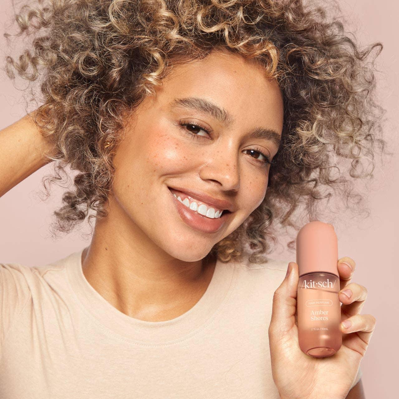 Smiling woman with curly hair holding Kitsch Amber Shores perfume, Come see us at Awaken, Franklin, Tennessee