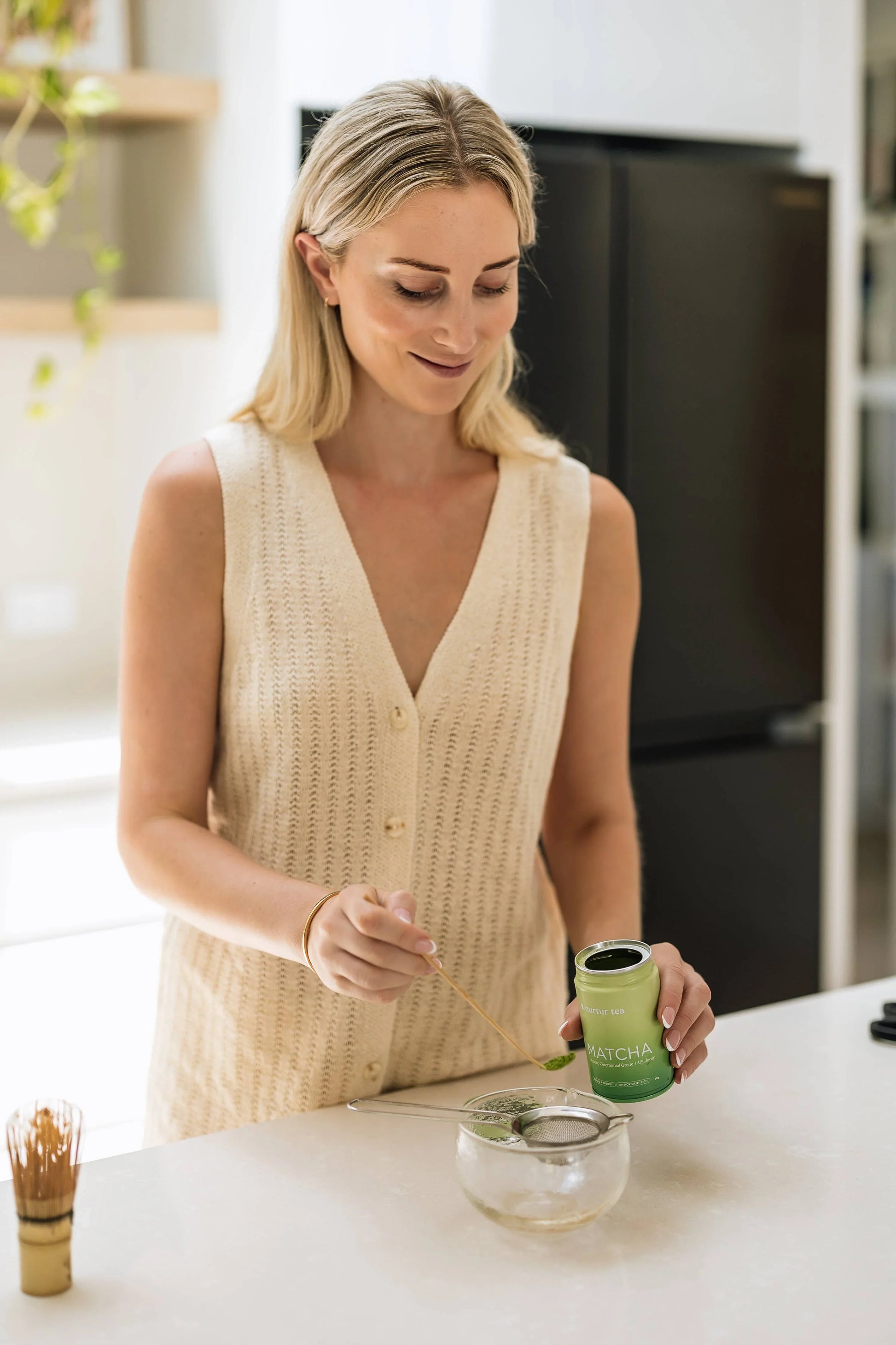 Woman in beige sleeveless top preparing matcha tea in kitchen. Come see us at Awaken, Franklin, Tennessee