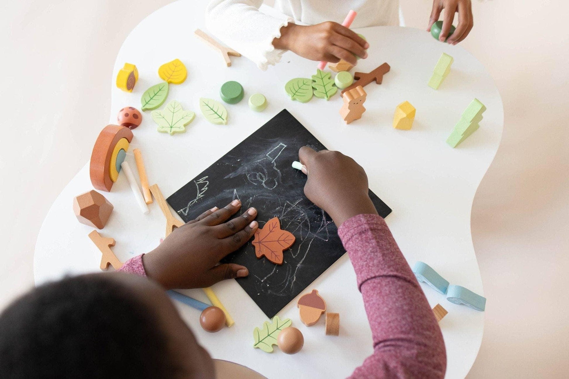 Children drawing with chalk and playing with wooden nature toys on white table. Come see us at Awaken, Franklin, Tennessee