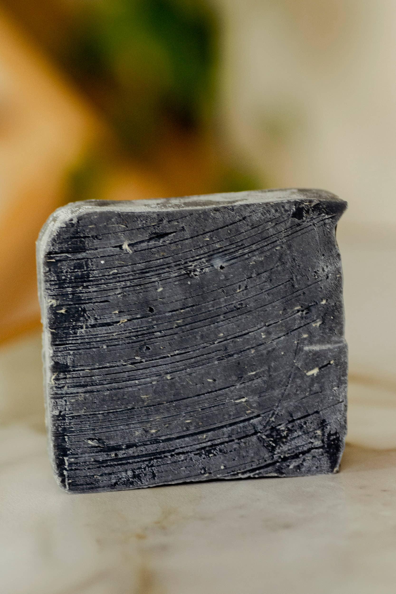 Close-up of black charcoal natural soap bar on marble surface with blurred plant in background. Come see us at Awaken, Franklin, Tennessee