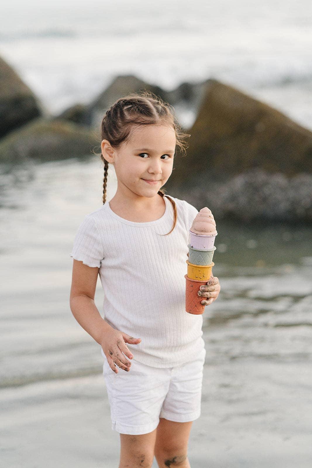Young girl in white outfit holding colorful sand cones by the water with rocks in background. Come see us at Awaken, Franklin, Tennessee