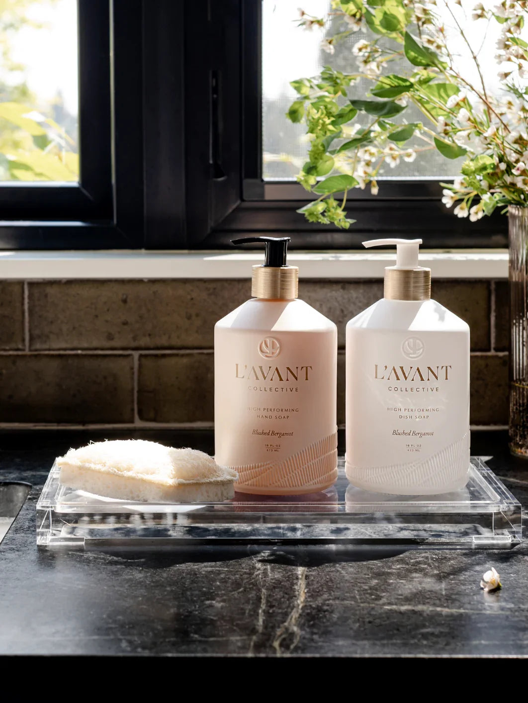 L'AVANT Collective hand soap and dish soap on a tray by a window with plants, sponge, and sunlight. Come see us at Awaken, Franklin, Tennessee.