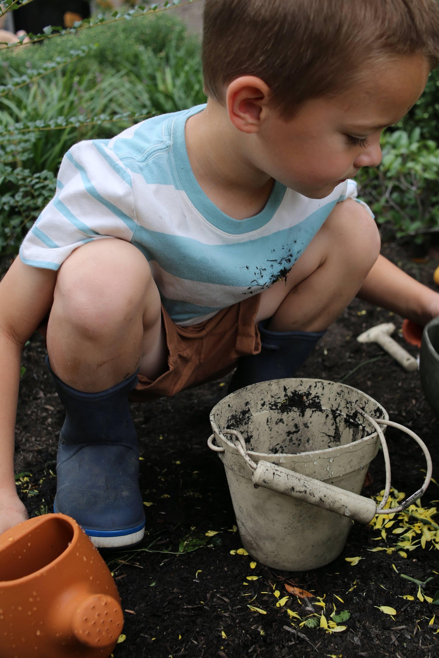 Young child in striped shirt and blue boots gardening with dirty white bucket and orange watering can, Come see us at Awaken, Franklin, Tennessee