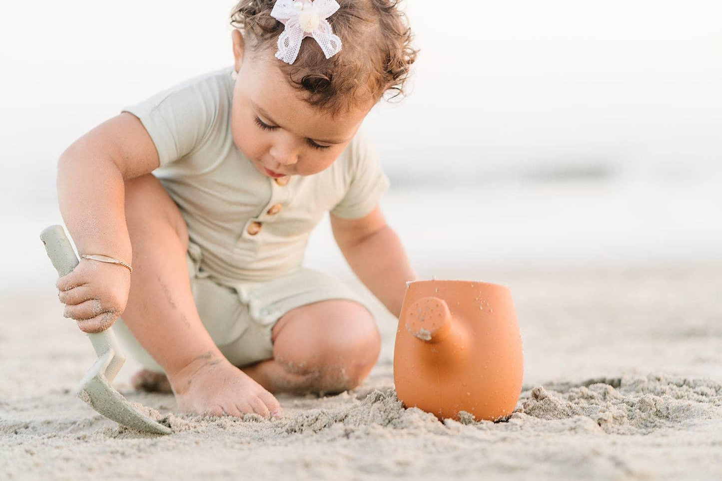 Toddler with curly hair playing in sand using a toy shovel and watering can. Come see us at Awaken, Franklin, Tennessee