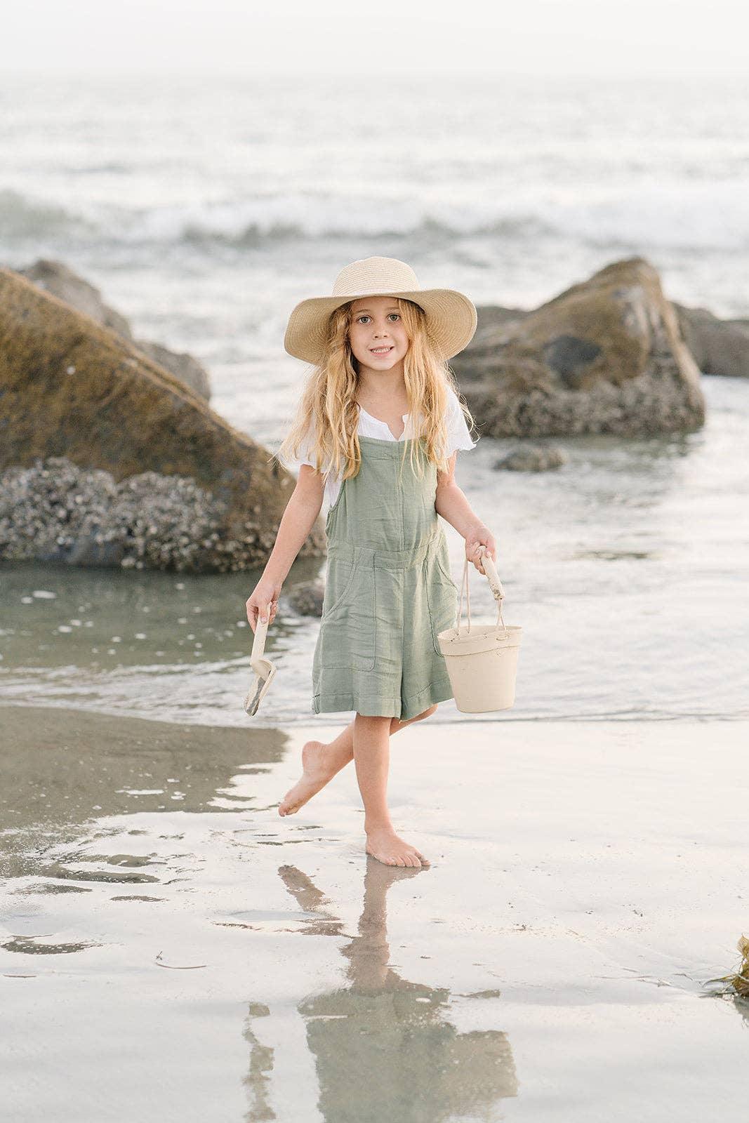 Young girl in sun hat and green dress holding bucket and shovel at beach with rocks. Come see us at Awaken, Franklin, Tennessee