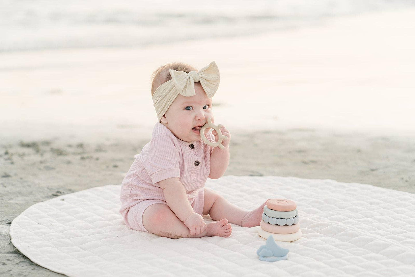 Baby in pink romper with cream bow headband sitting on quilted mat playing with teething toys at beach. Come see us at Awaken, Franklin, Tennessee
