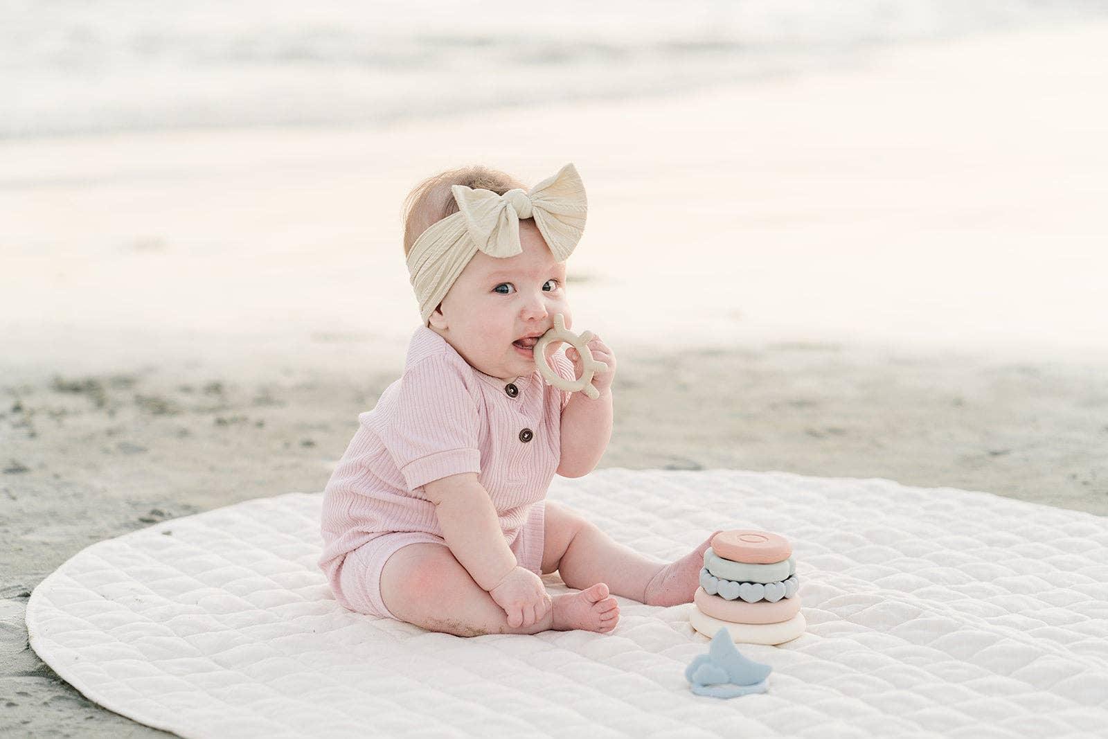 Baby in pink romper with cream bow headband sitting on quilted mat playing with teething toys at beach. Come see us at Awaken, Franklin, Tennessee