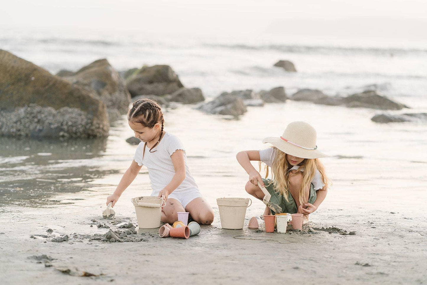 Two children playing with sand and buckets on a beach near rocks and ocean waves. Come see us at Awaken, Franklin, Tennessee