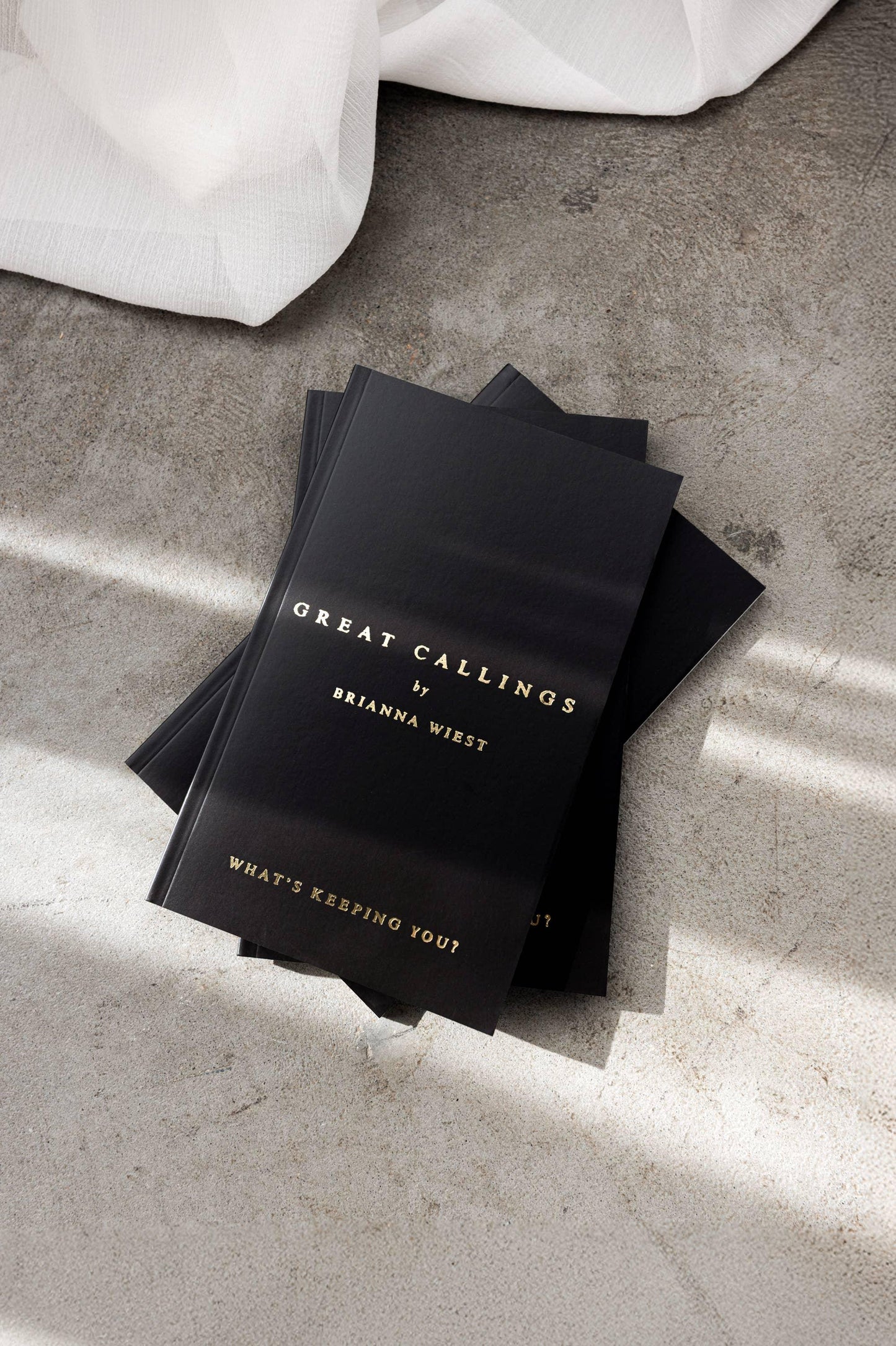 Stack of black books titled Great Callings by Brianna Wiest on concrete floor with fabric, Come see us at Awaken, Franklin, Tennessee