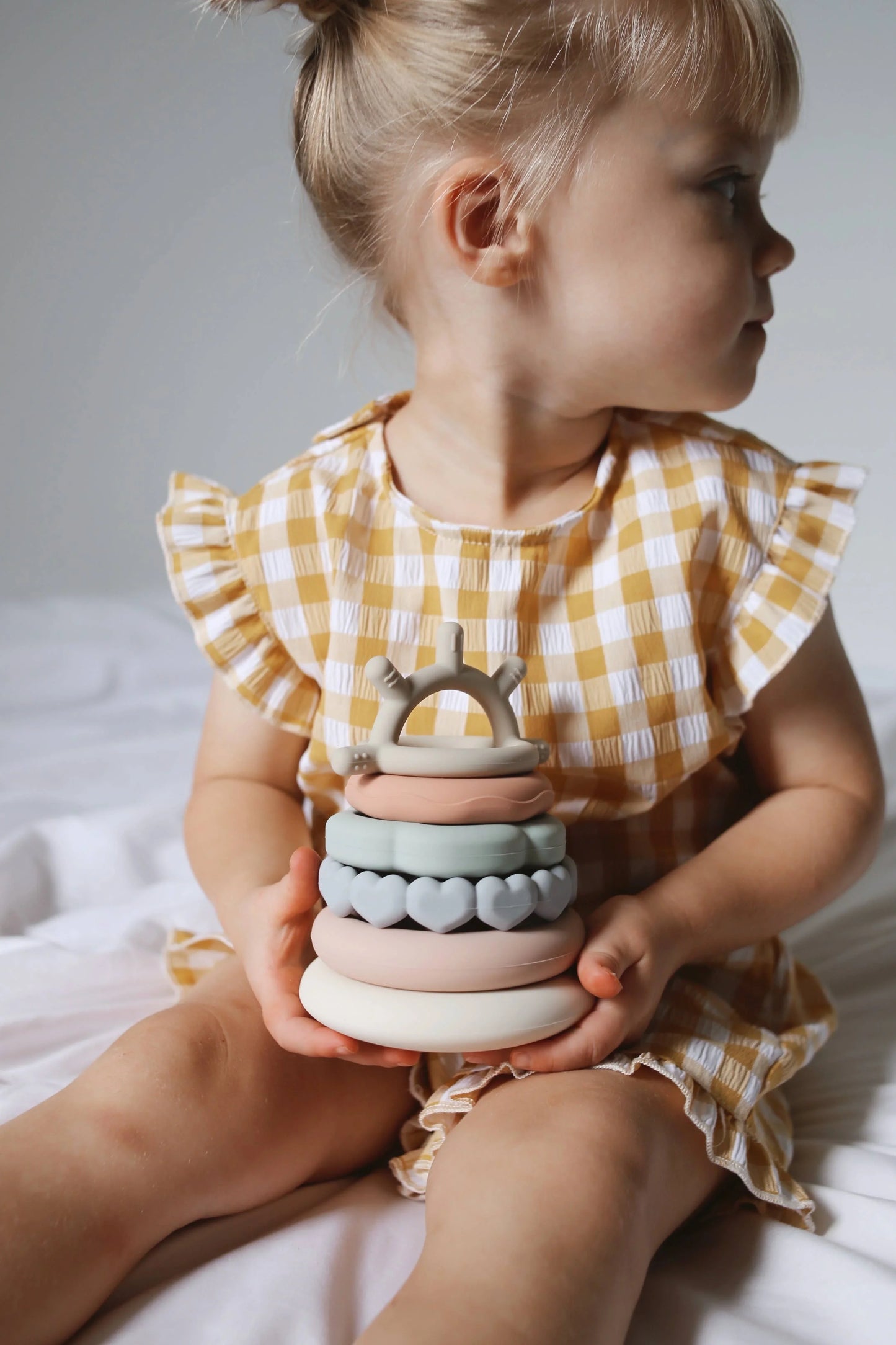 Toddler in yellow gingham dress holding pastel silicone stacking rings toy on bed. Come see us at Awaken, Franklin, Tennessee