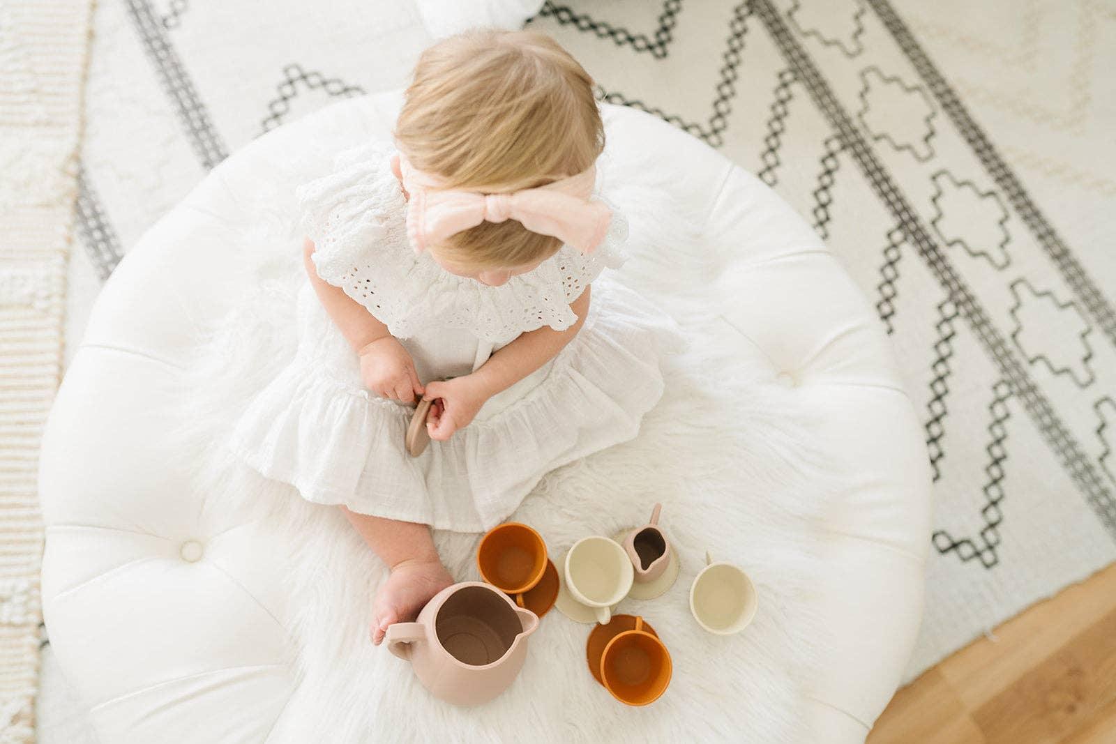 Baby girl in white dress with pink bow headband playing tea set on ottoman, Come see us at Awaken, Franklin, Tennessee