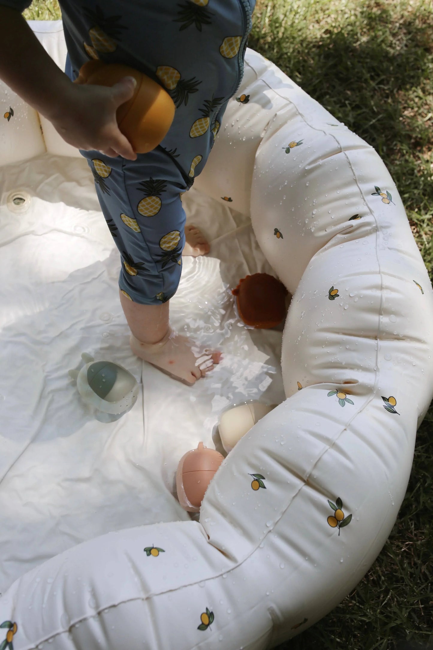 Toddler in pineapple-print swimsuit playing in inflatable pool outdoors with toys. Come see us at Awaken, Franklin, Tennessee