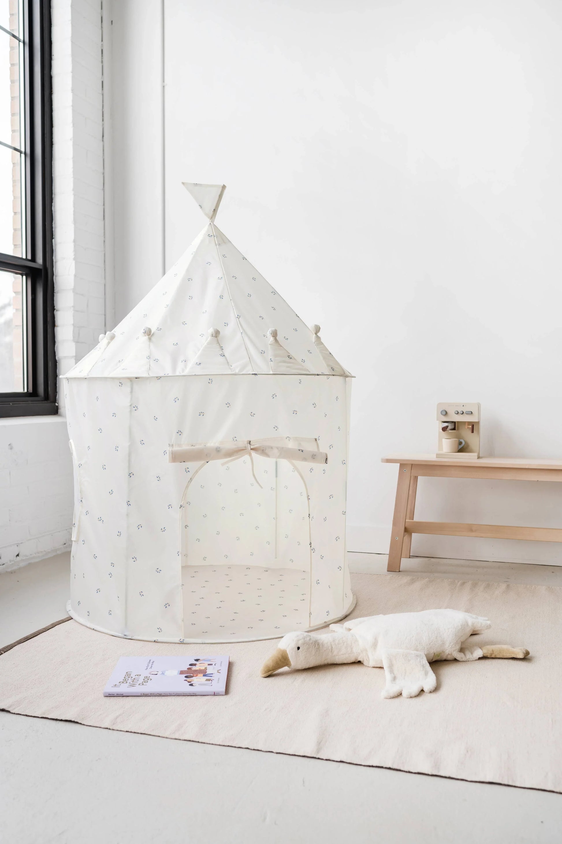 White play tent with small blue floral patterns, plush bird toy, book, and wooden bench in minimalist room. Come see us at Awaken, Franklin, Tennessee
