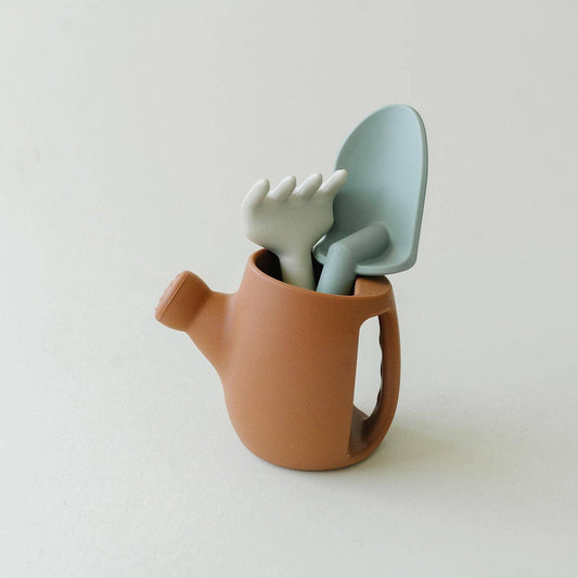 Clay brown watering can holding a green silicone rake and light blue shovel on white surface. Come see us at Awaken, Franklin, Tennessee
