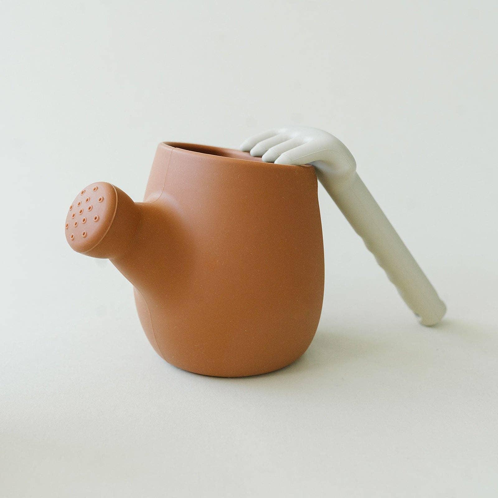 Modern brown watering can with gray hand-shaped handle on white background. Come see us at Awaken, Franklin, Tennessee