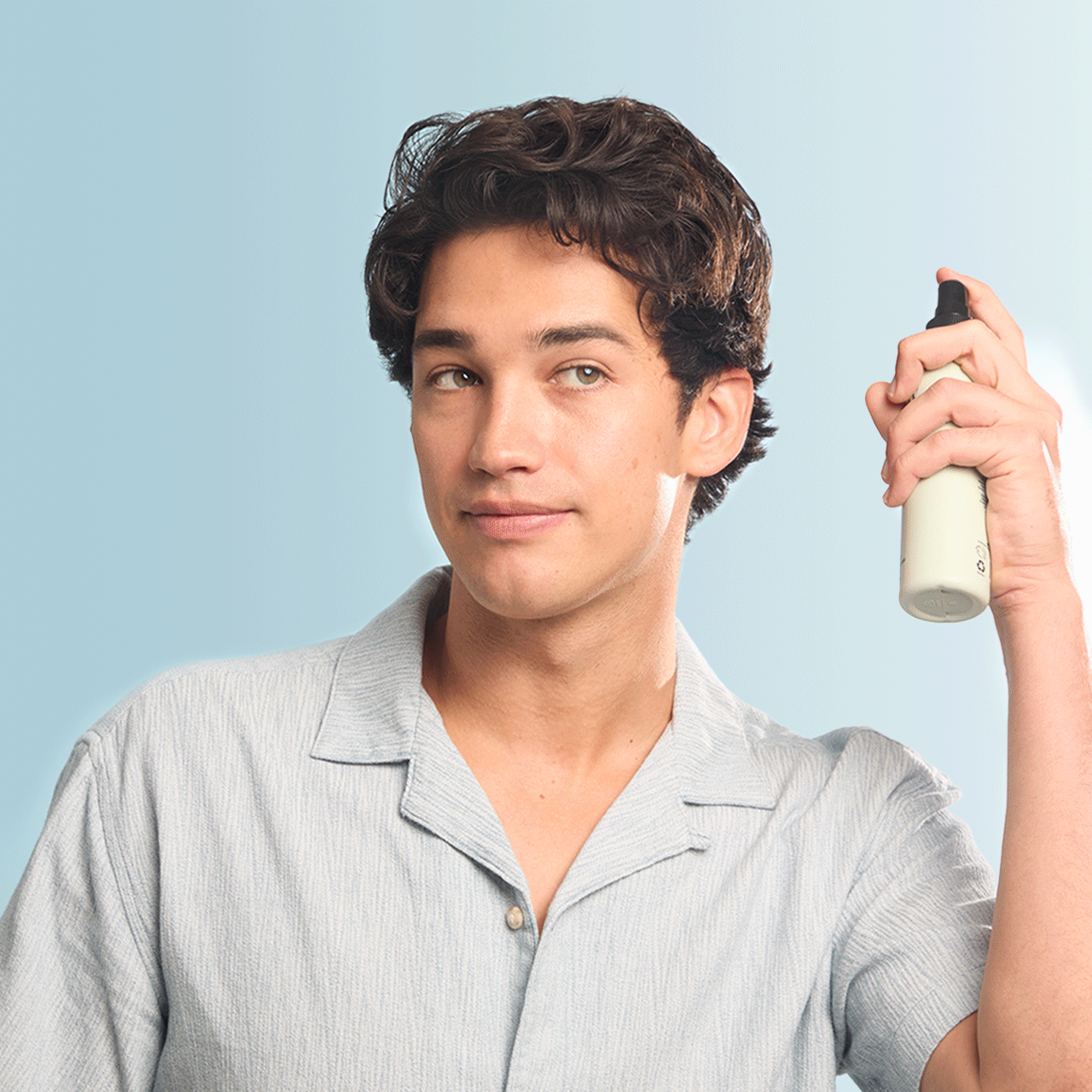 Young man spraying hair product with light blue shirt on plain background. Come see us at Awaken, Franklin, Tennessee