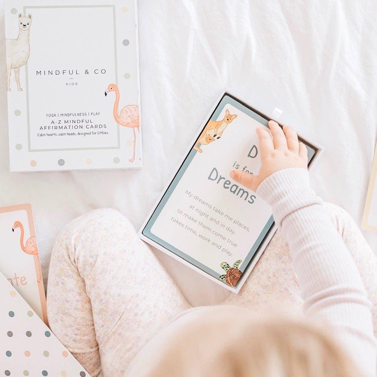 Child holding mindfulness affirmation card 'Dreams' from Mindful & Co Kids set on white bed. Come see us at Awaken, Franklin, Tennessee
