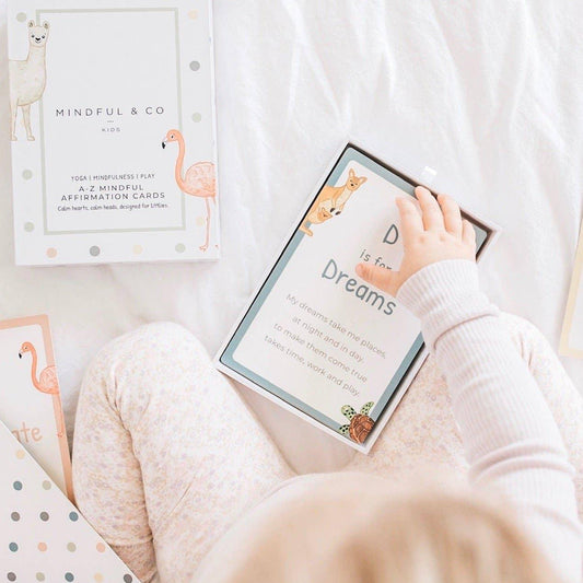 Child holding mindfulness affirmation card 'Dreams' from Mindful & Co Kids set on white bed. Come see us at Awaken, Franklin, Tennessee