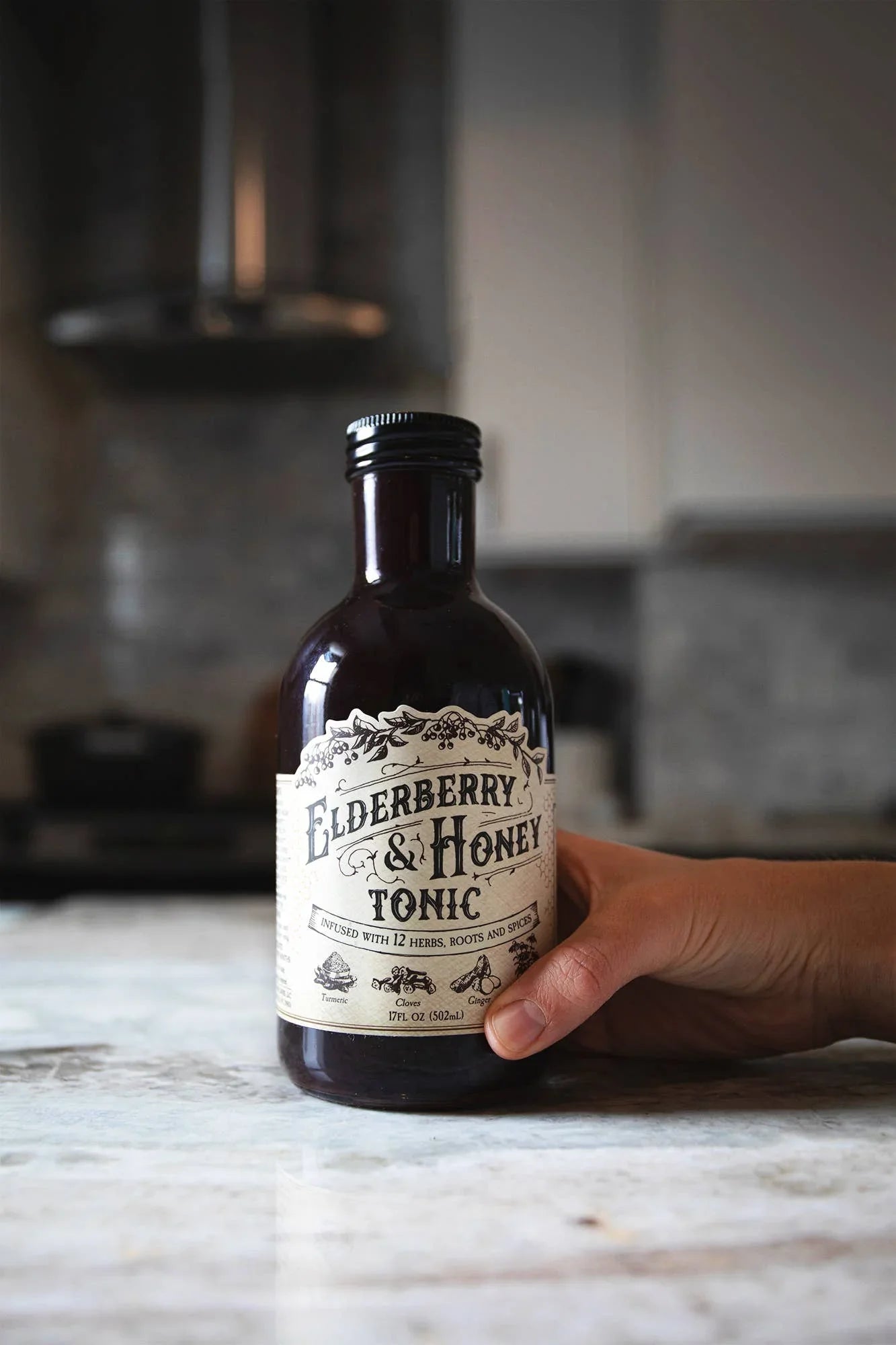 Hand holding a dark bottle of elderberry & honey tonic on marble kitchen counter, Come see us at Awaken, Franklin, Tennessee