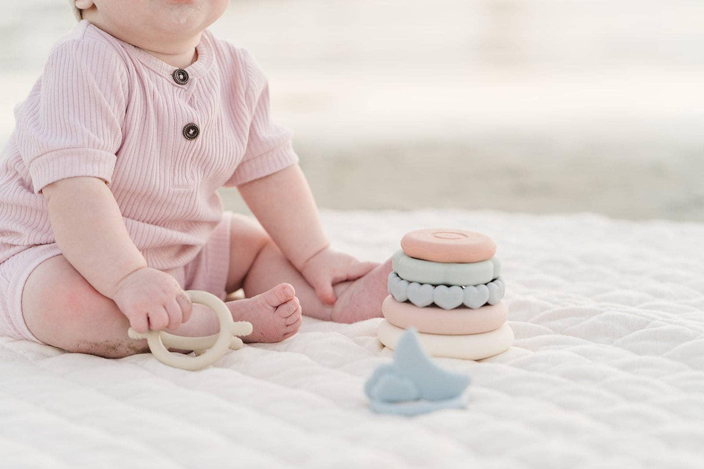 Baby in pink onesie playing with pastel stacking rings and teether on white quilt. Come see us at Awaken, Franklin, Tennessee