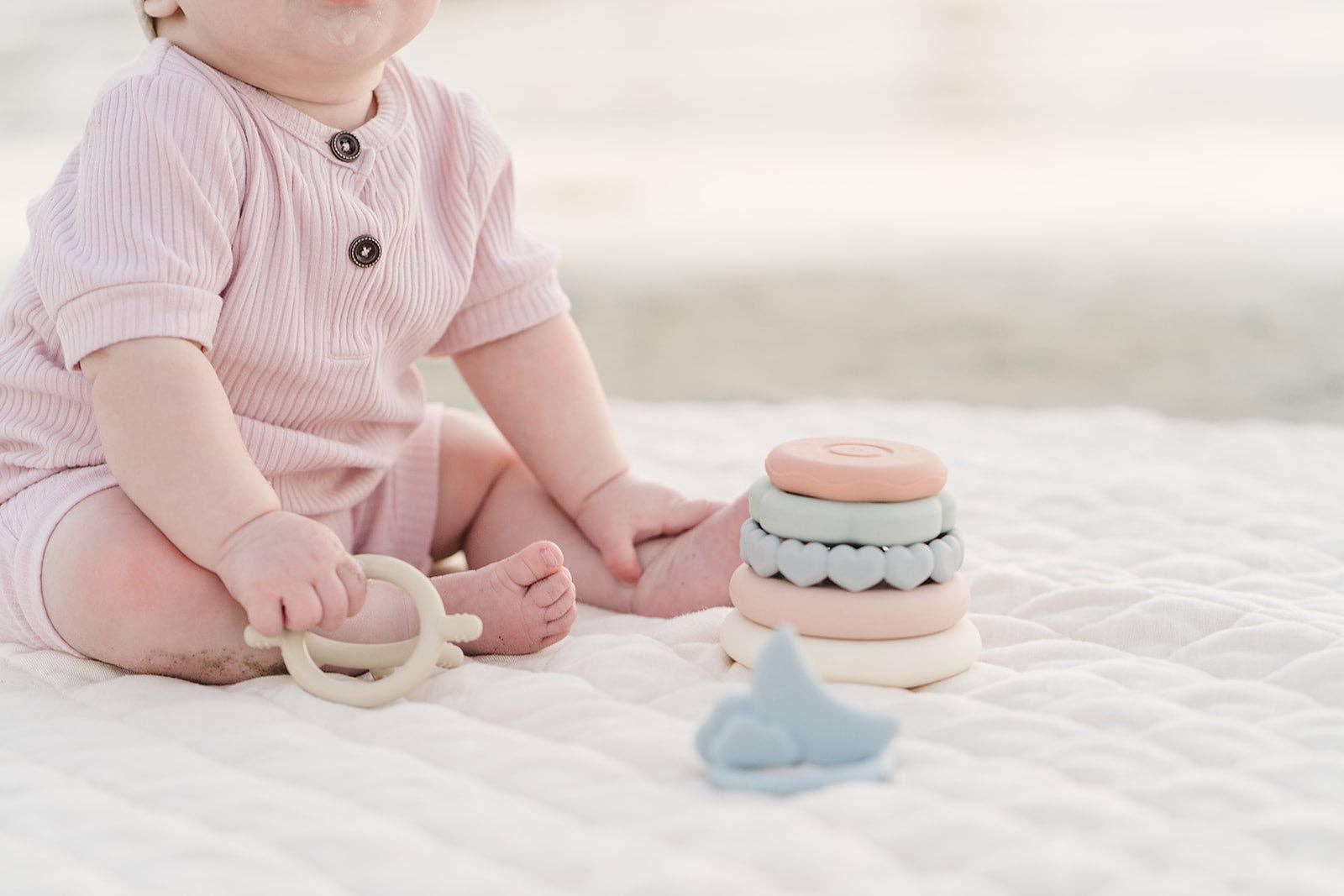 Baby in pink onesie playing with pastel stacking rings and teether on white quilt. Come see us at Awaken, Franklin, Tennessee
