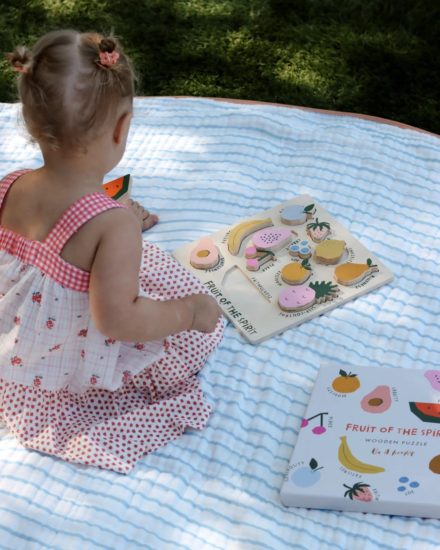 Toddler in floral dress playing with Fruit of the Spirit wooden puzzle outdoors on blanket. Come see us at Awaken, Franklin, Tennessee