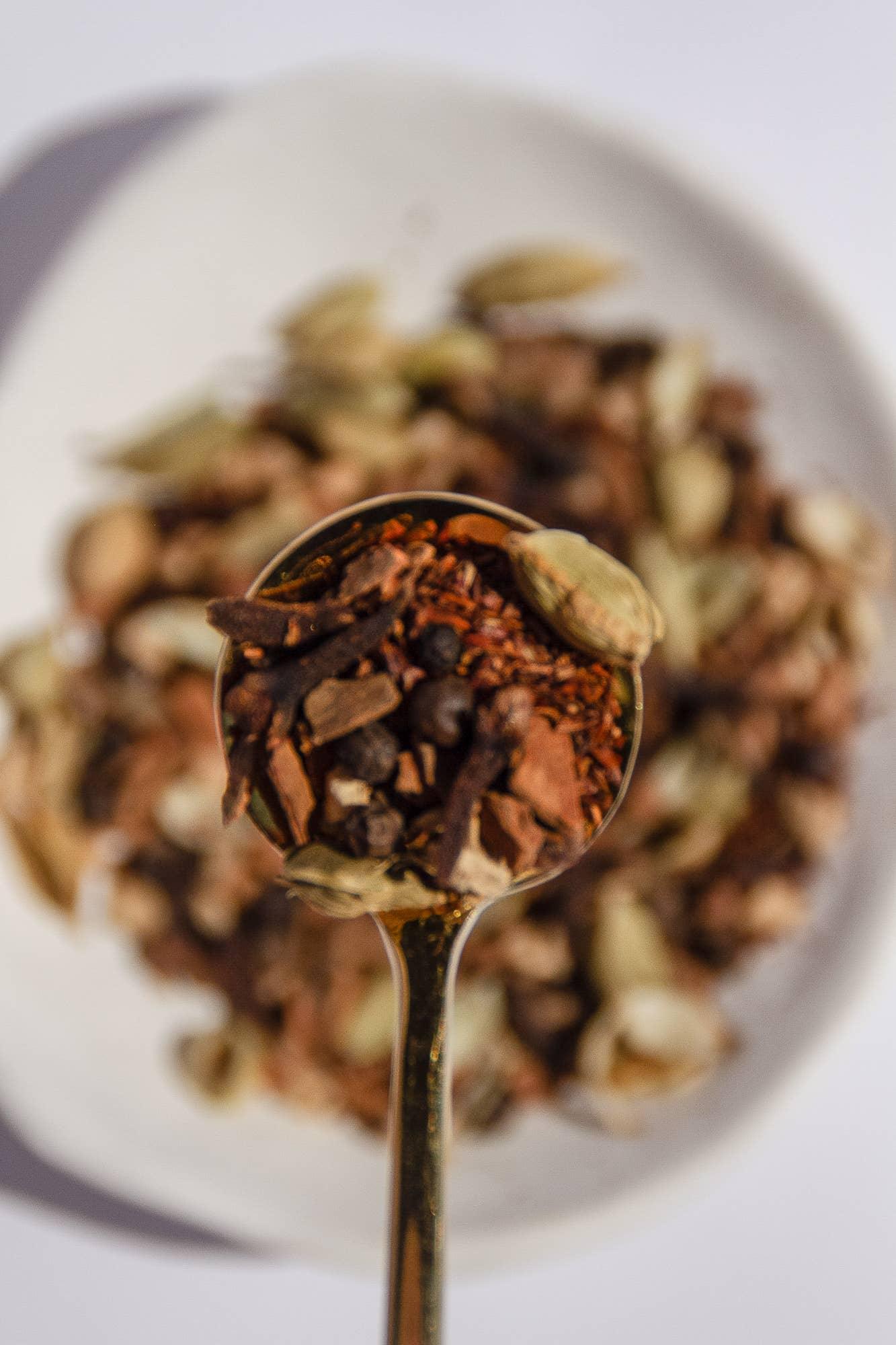 Close-up of a spoon with mixed loose spices including cloves and cardamom on a white plate background. Come see us at Awaken, Franklin, Tennessee