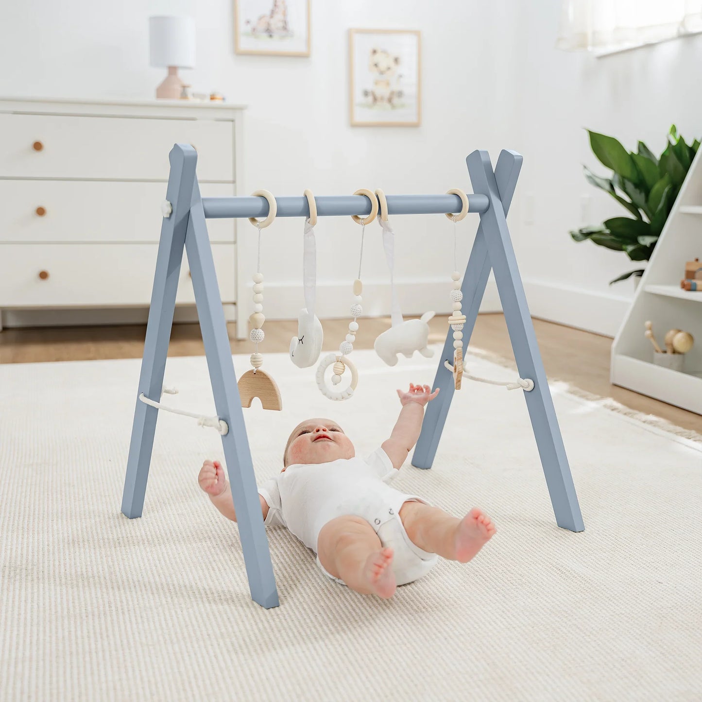 Baby lying on carpet reaching toward blue wooden baby gym with hanging toys in bright nursery Come see us at Awaken, Franklin, Tennessee