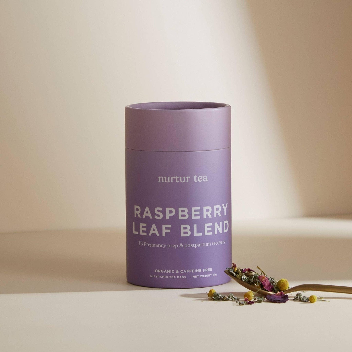 Purple Nurtur Tea Raspberry Leaf Blend container with dried herbs on spoon, soft beige background. Come see us at Awaken, Franklin, Tennessee