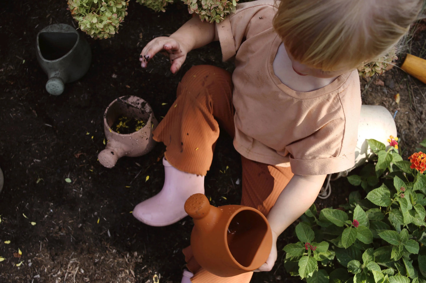 Toddler in beige shirt and rust pants holding clay watering can, gardening outdoors with green plants. Come see us at Awaken, Franklin, Tennessee