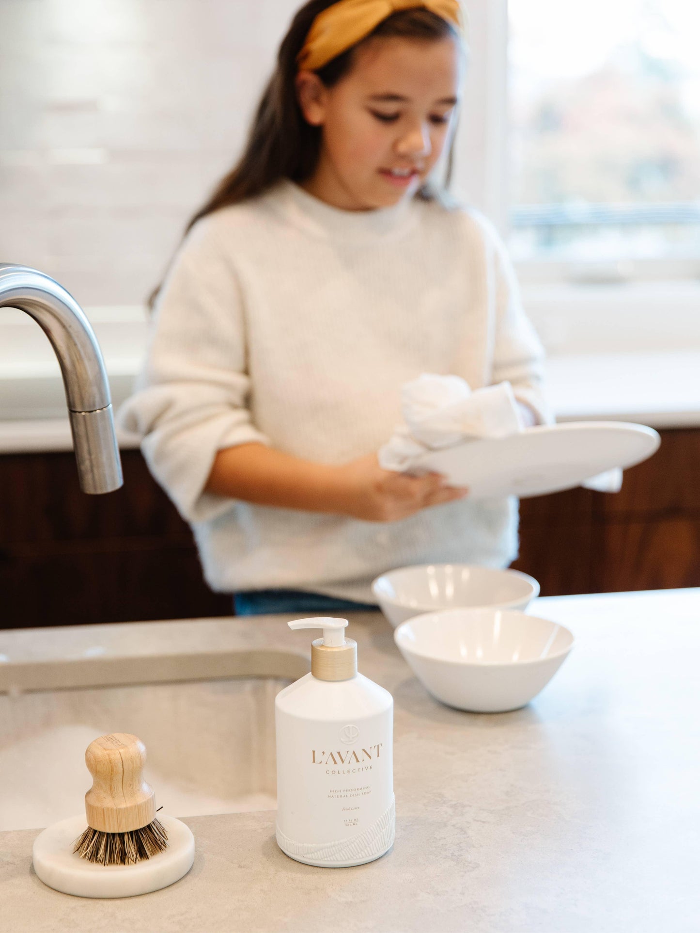 Girl cleaning dishes in a bright kitchen with L'AVANT soap pump and brush on counter. Come see us at Awaken, Franklin, Tennessee.