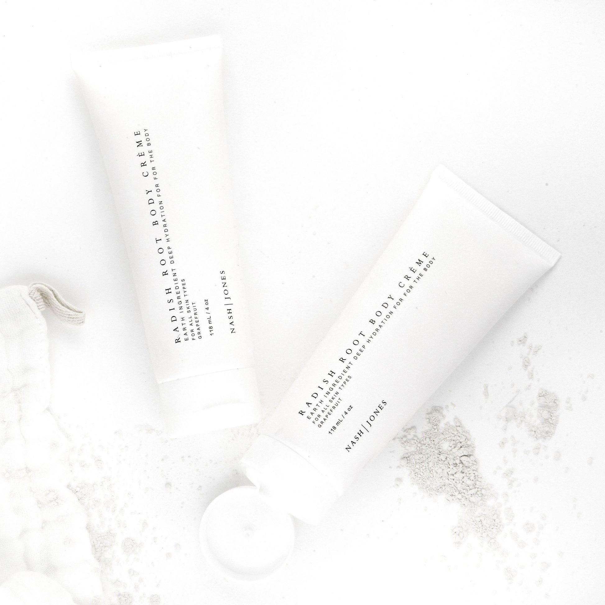 Two tubes of Nash Jones Radish Root Body Crème with grapefruit scent on a white background with powder spills. Come see us at Awaken, Franklin, Tennessee