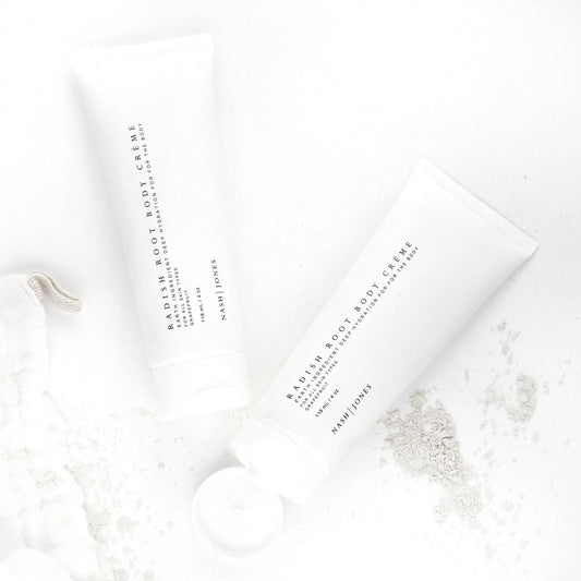 Two tubes of Nash Jones Radish Root Body Crème with grapefruit scent on a white background with powder spills. Come see us at Awaken, Franklin, Tennessee