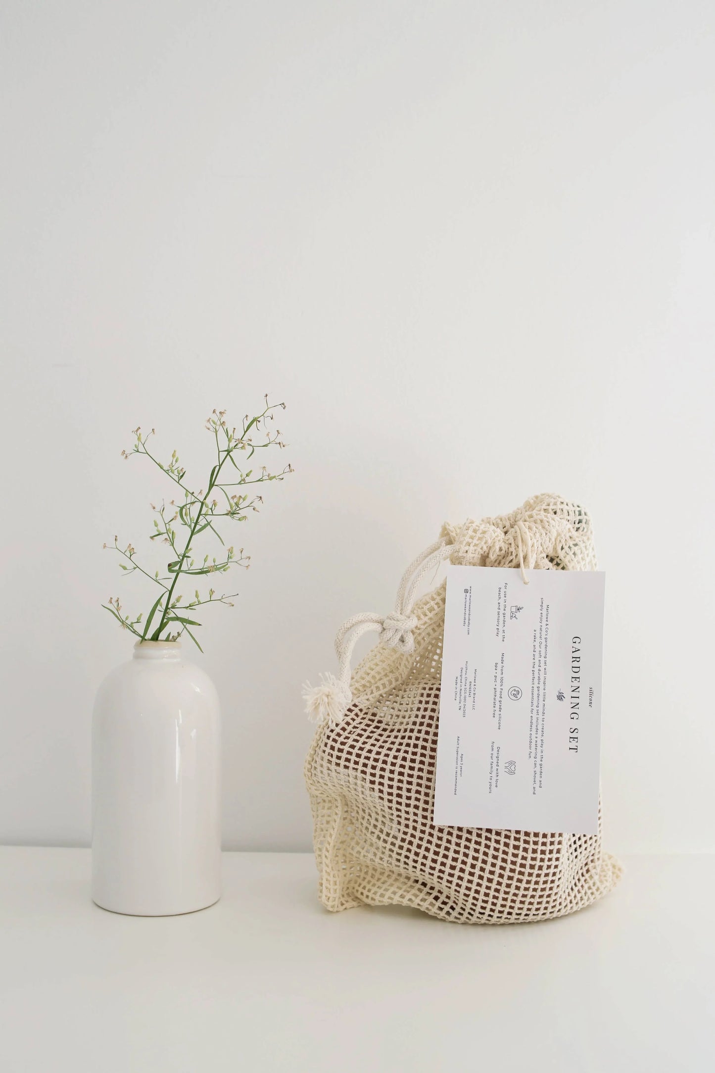 Minimalist white vase with delicate green sprig beside a beige mesh bag labeled gardening set on white table. Come see us at Awaken, Franklin, Tennessee