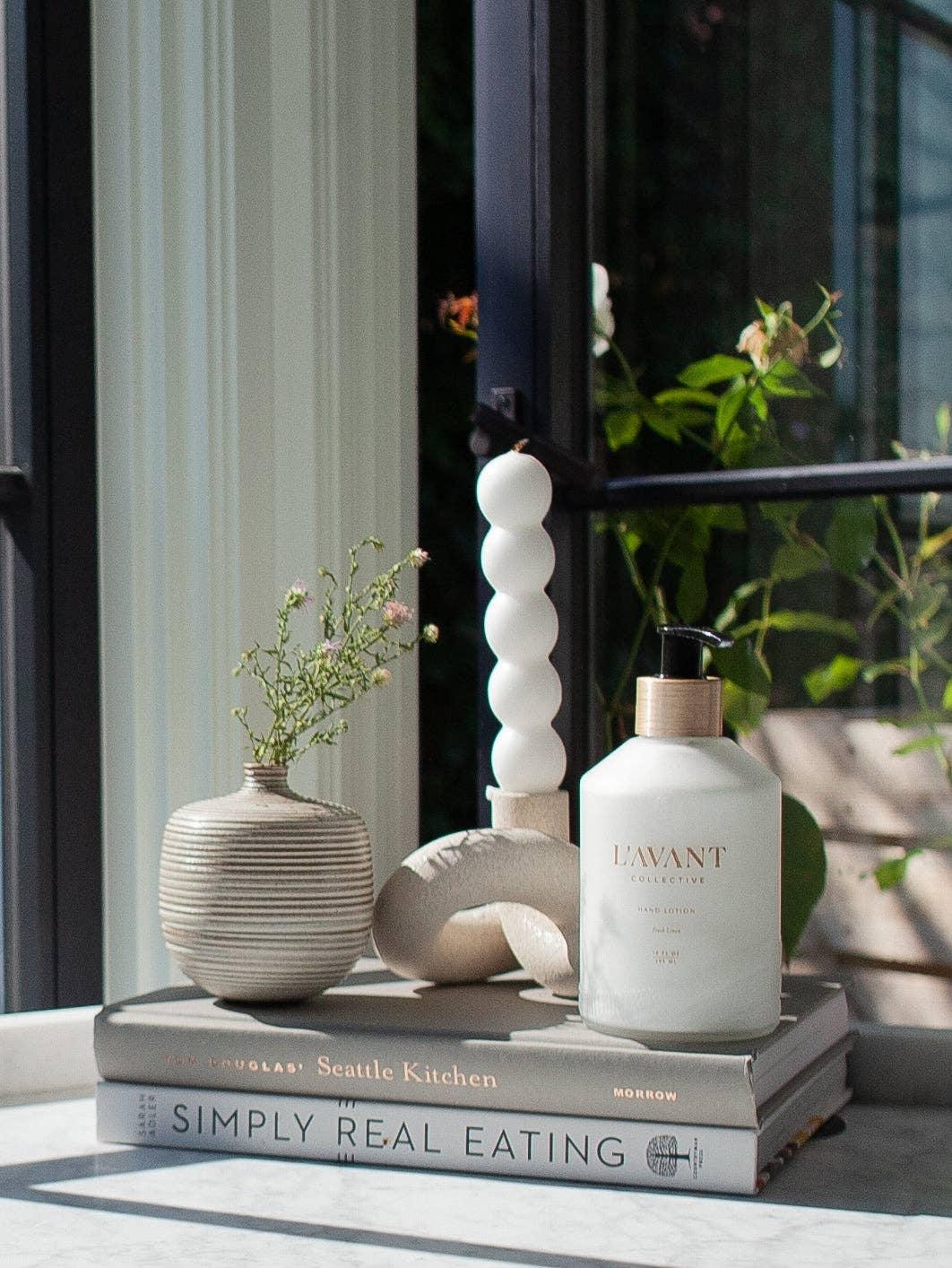 L'Avant Collective hand lotion on books beside a small striped vase, bright window; Come see us at Awaken, Franklin, Tennessee