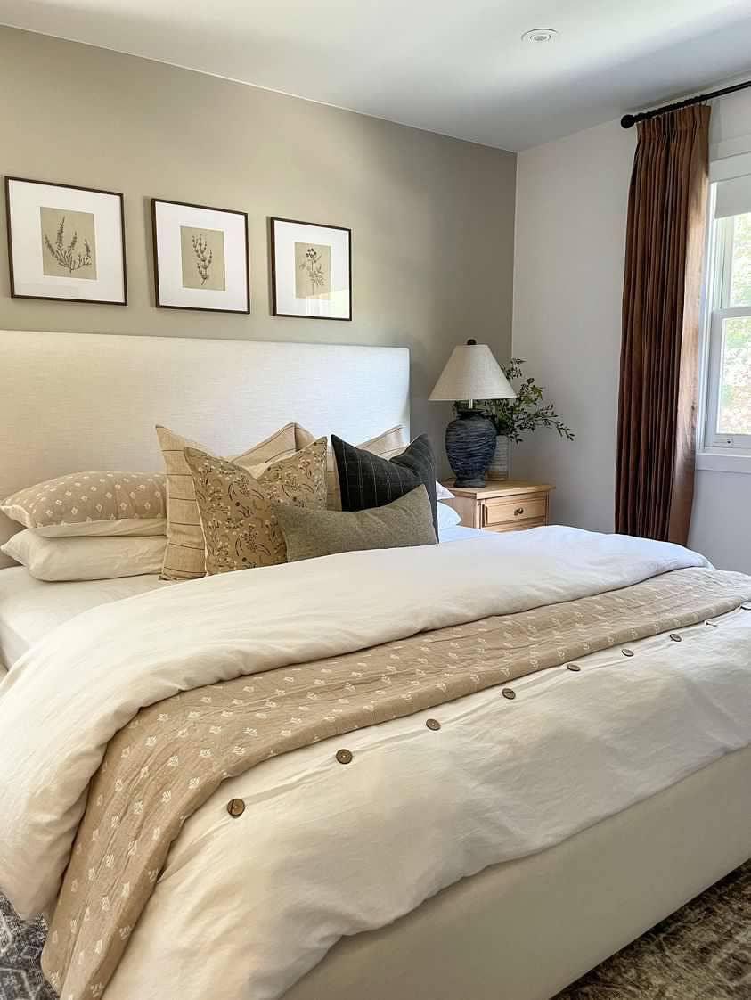 Cozy bedroom with beige bedding, assorted pillows, nightstand with lamp, and wall art. Come see us at Awaken, Franklin, Tennessee