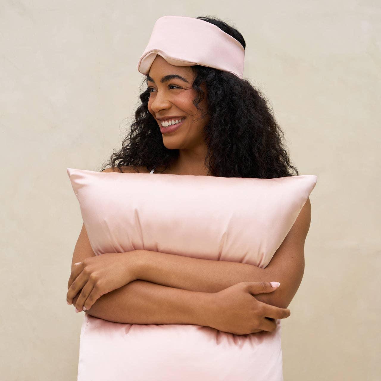 Smiling woman with curly hair hugging a pale pink satin pillow and wearing a matching sleep mask, neutral background. Come see us at Awaken, Franklin, Tennessee