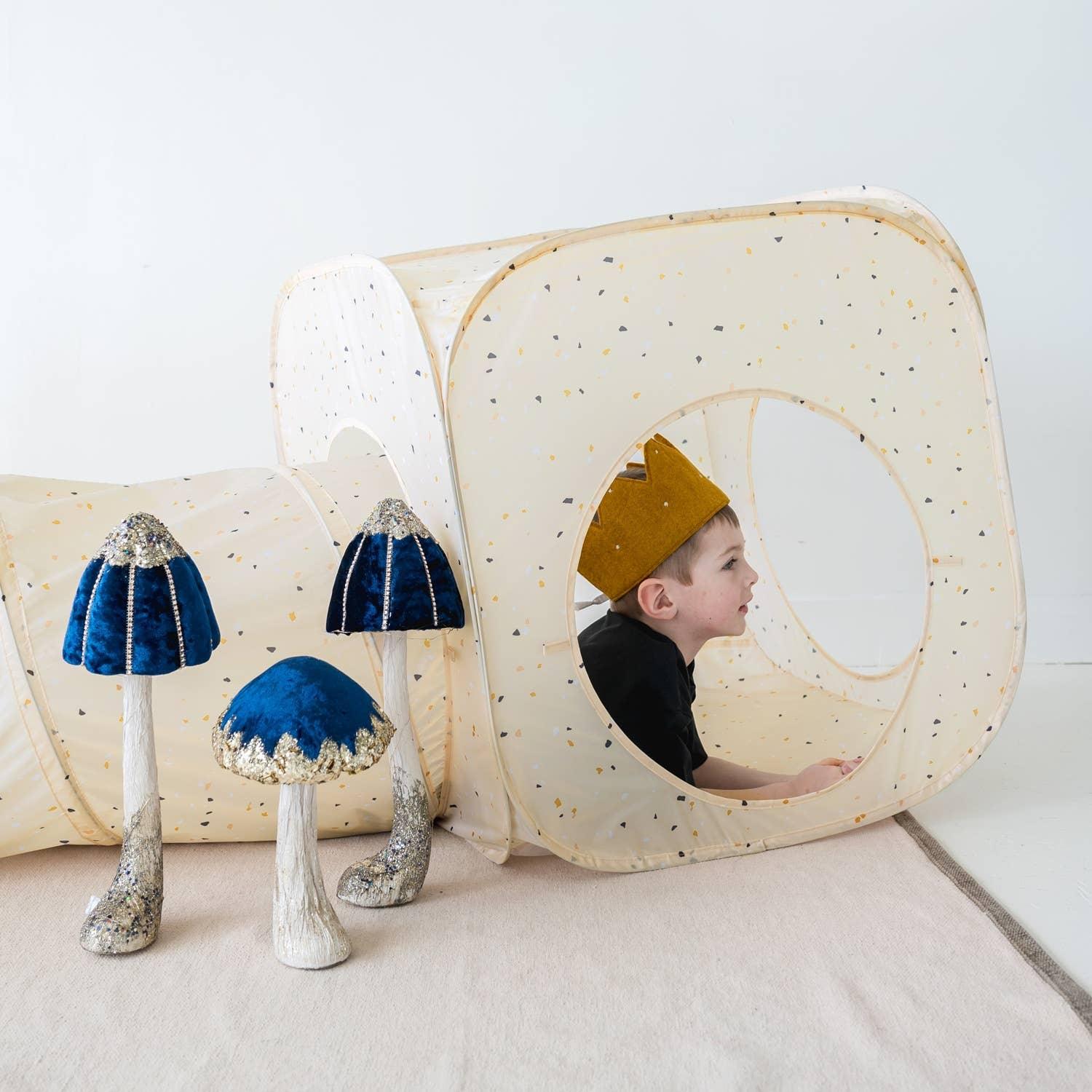 Child wearing crown playing inside beige speckled fabric tunnel with decorative blue velvet mushrooms. Come see us at Awaken, Franklin, Tennessee