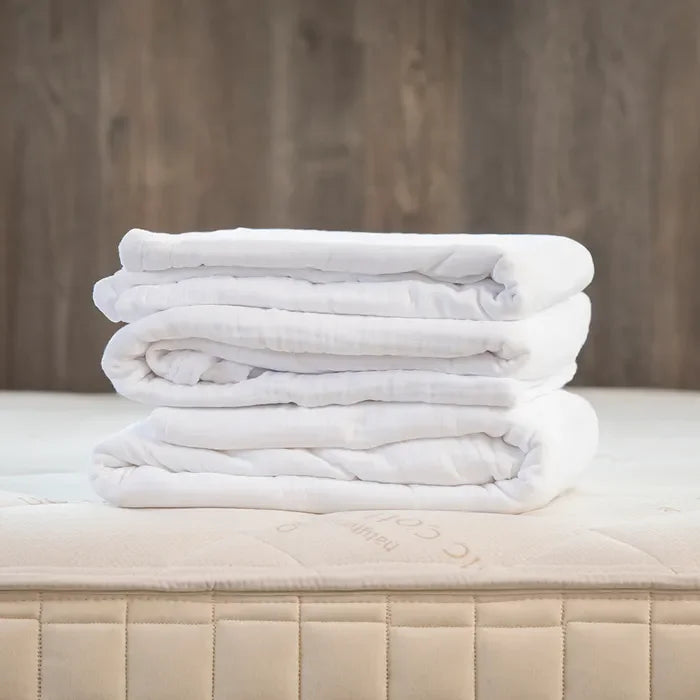 White muslin adult blanket neatly folded on a mattress, bedding display. Come see us at Awaken, Franklin, Tennessee.