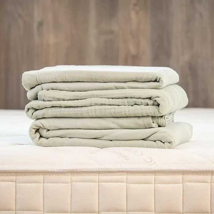 Sage green muslin adult blanket folded on a mattress, bedding display. Come see us at Awaken, Franklin, Tennessee.