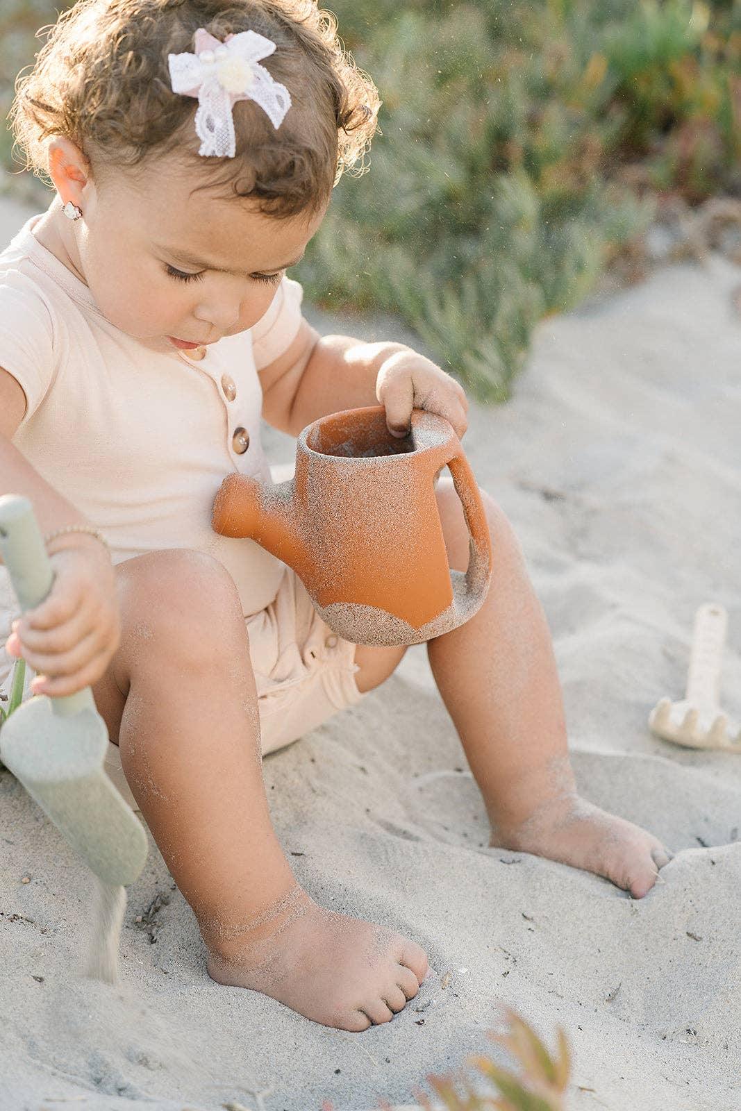 Toddler girl playing with a sand shovel and orange watering can outdoors. Come see us at Awaken, Franklin, Tennessee