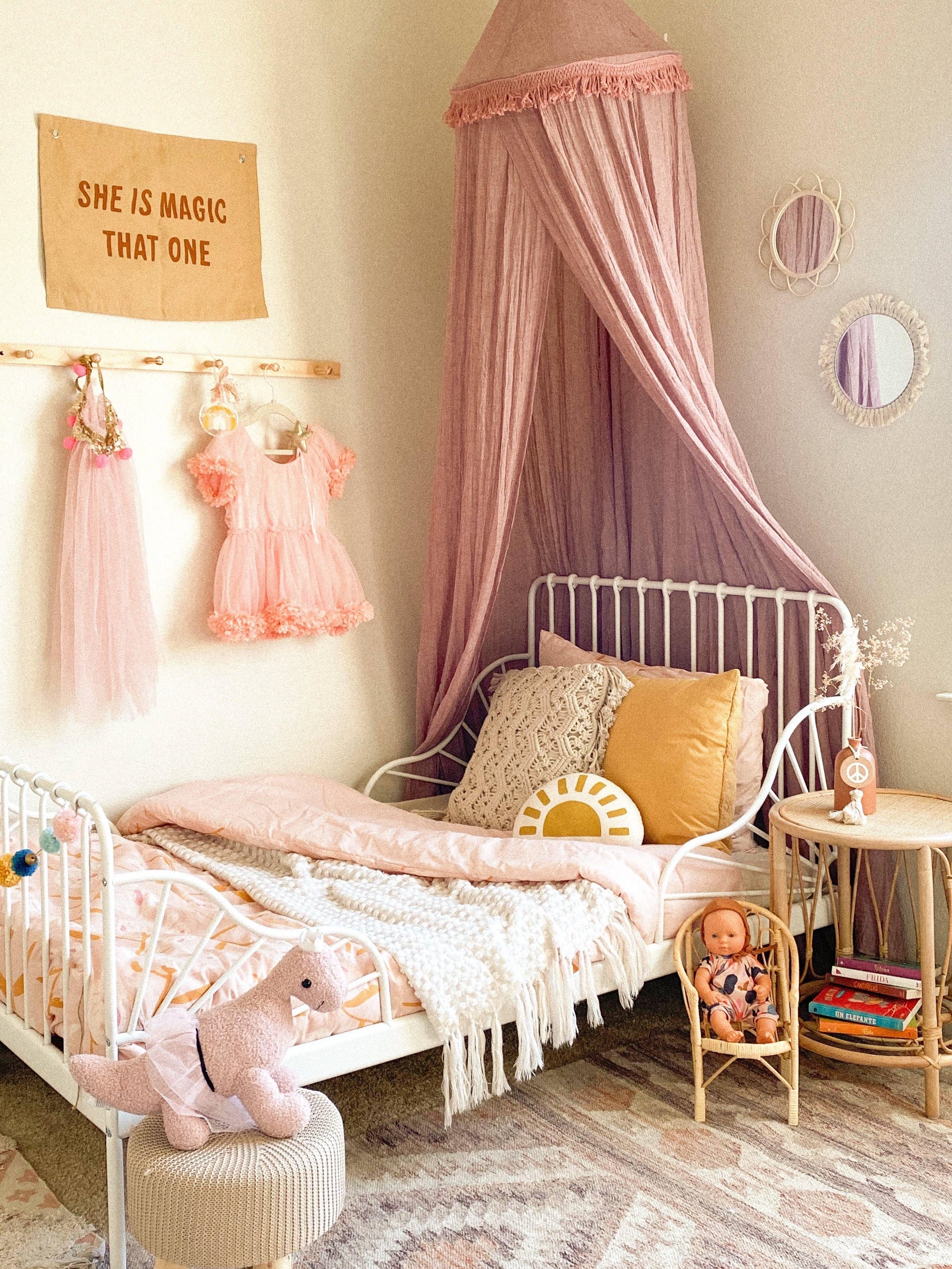 Boho girls' bedroom with pink canopy, plush dinosaur, dolls, and playful decor. Come see us at Awaken, Franklin, Tennessee.