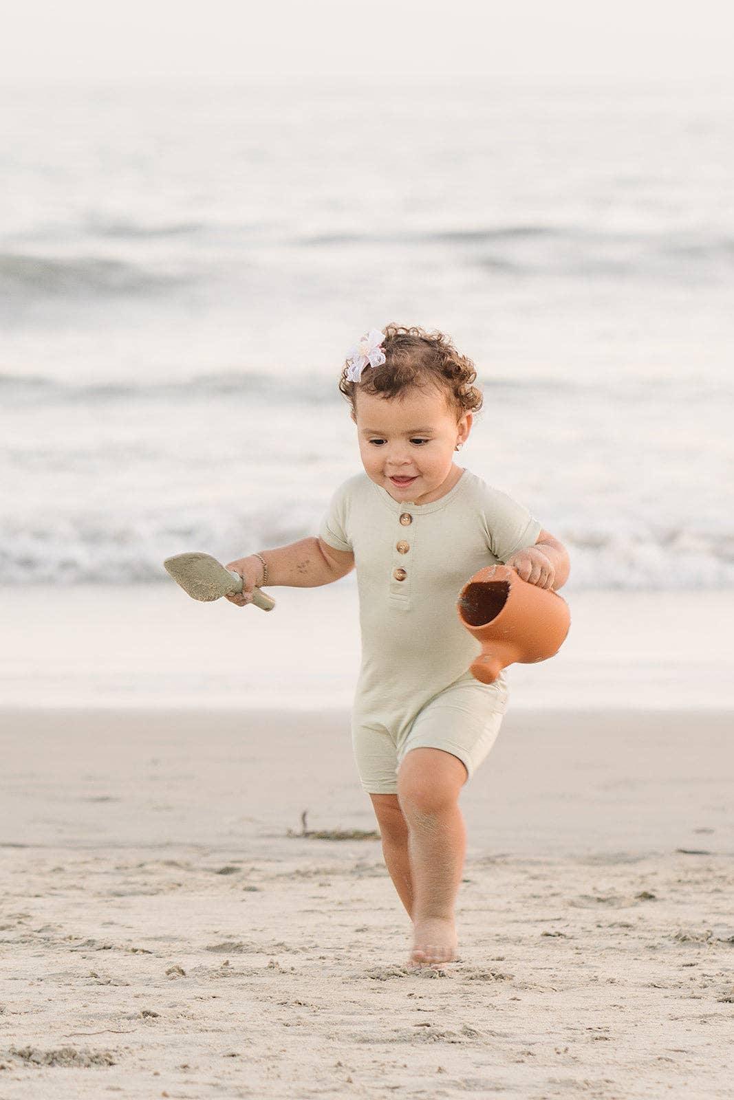 Toddler in beige romper playing with clay pot and shovel on sandy beach at sunset. Come see us at Awaken, Franklin, Tennessee