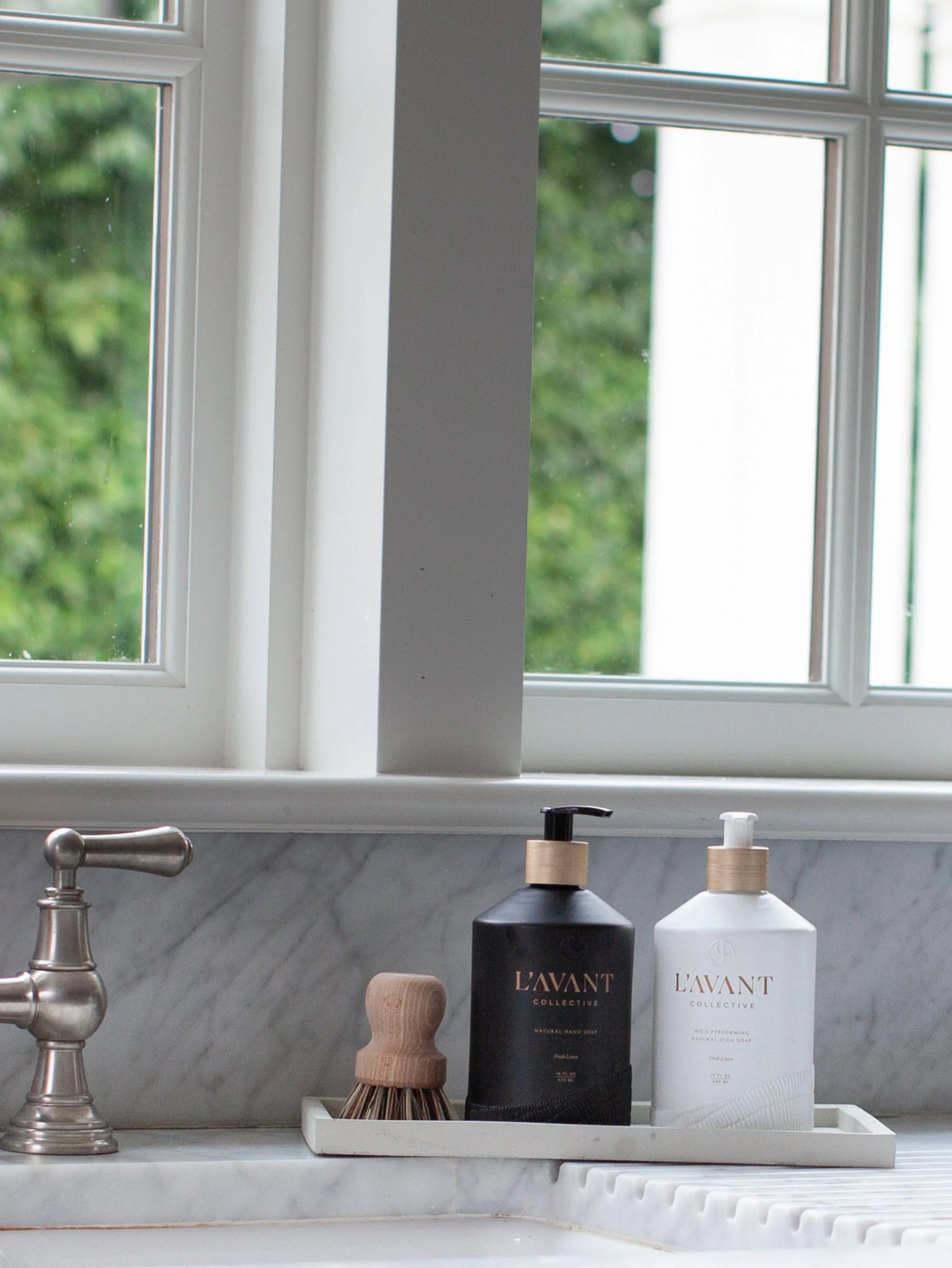 L'AVANT Collective hand soaps on marble sink with wooden brush, kitchen window view. Come see us at Awaken, Franklin, Tennessee.