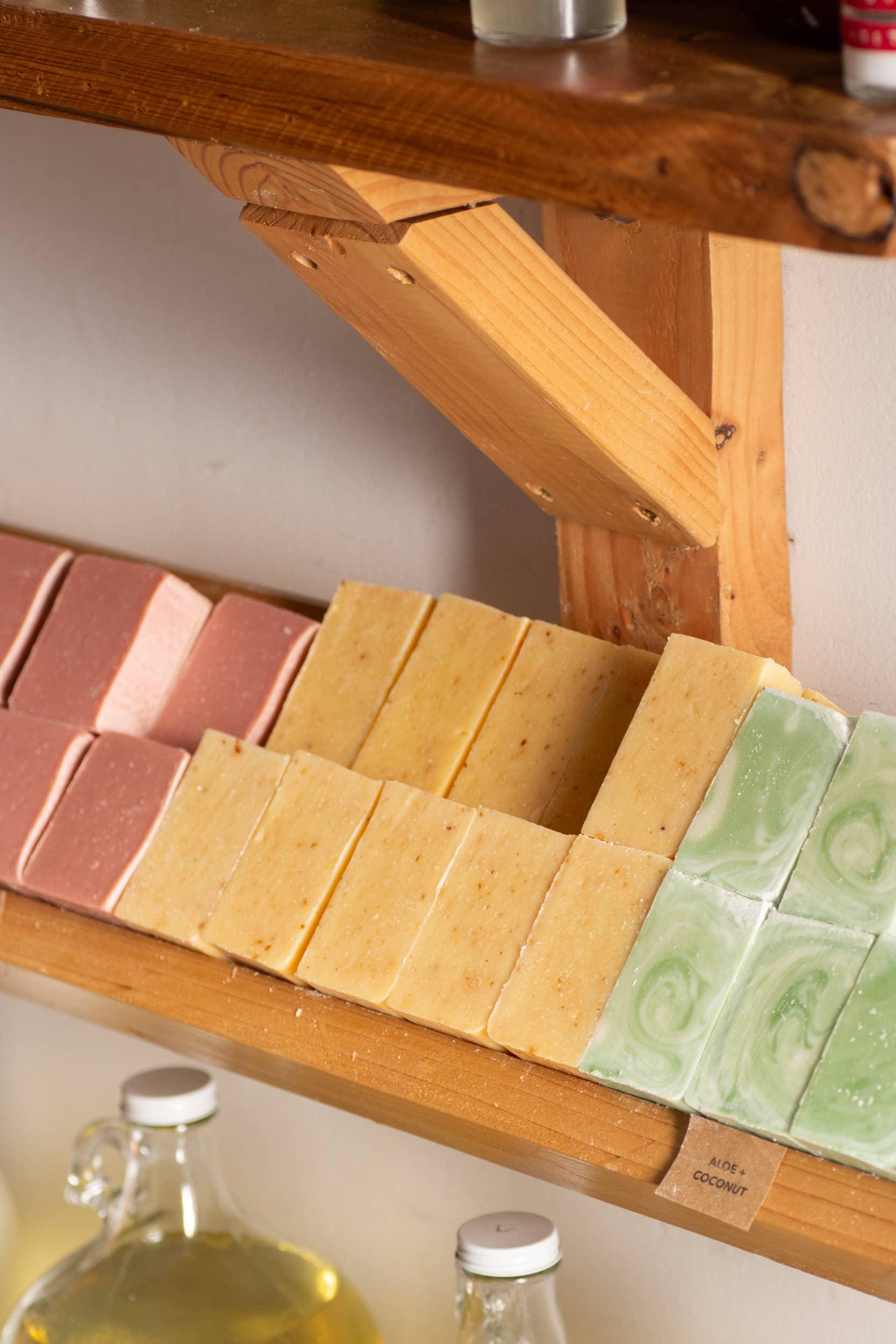 Handmade soap bars in pink, beige, and green aloe coconut on wooden shelf with glass bottles below. Come see us at Awaken, Franklin, Tennessee