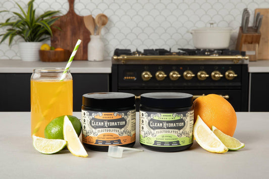 Two Clean Hydration electrolyte supplement containers with orange and lemon-lime flavors, fresh citrus fruits, and a glass of juice on kitchen counter. Come see us at Awaken, Franklin, Tennessee
