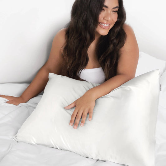 Smiling woman with long dark hair holding a white silk pillow on white bed linens, Come see us at Awaken, Franklin, Tennessee