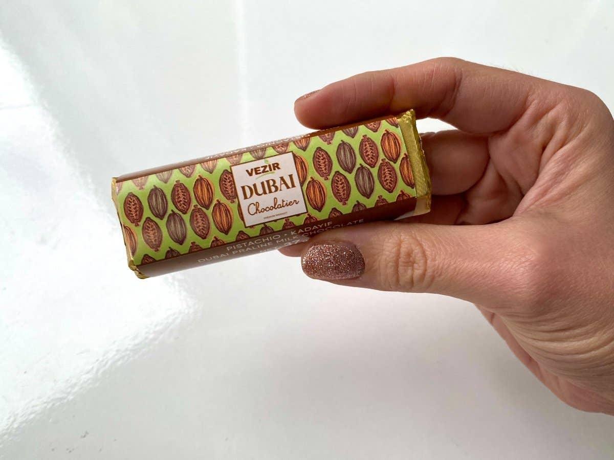 Hand holding Vezir Dubai Chocolatier pistachio kadayif praline chocolate bar, bright background, Come see us at Awaken, Franklin, Tennessee