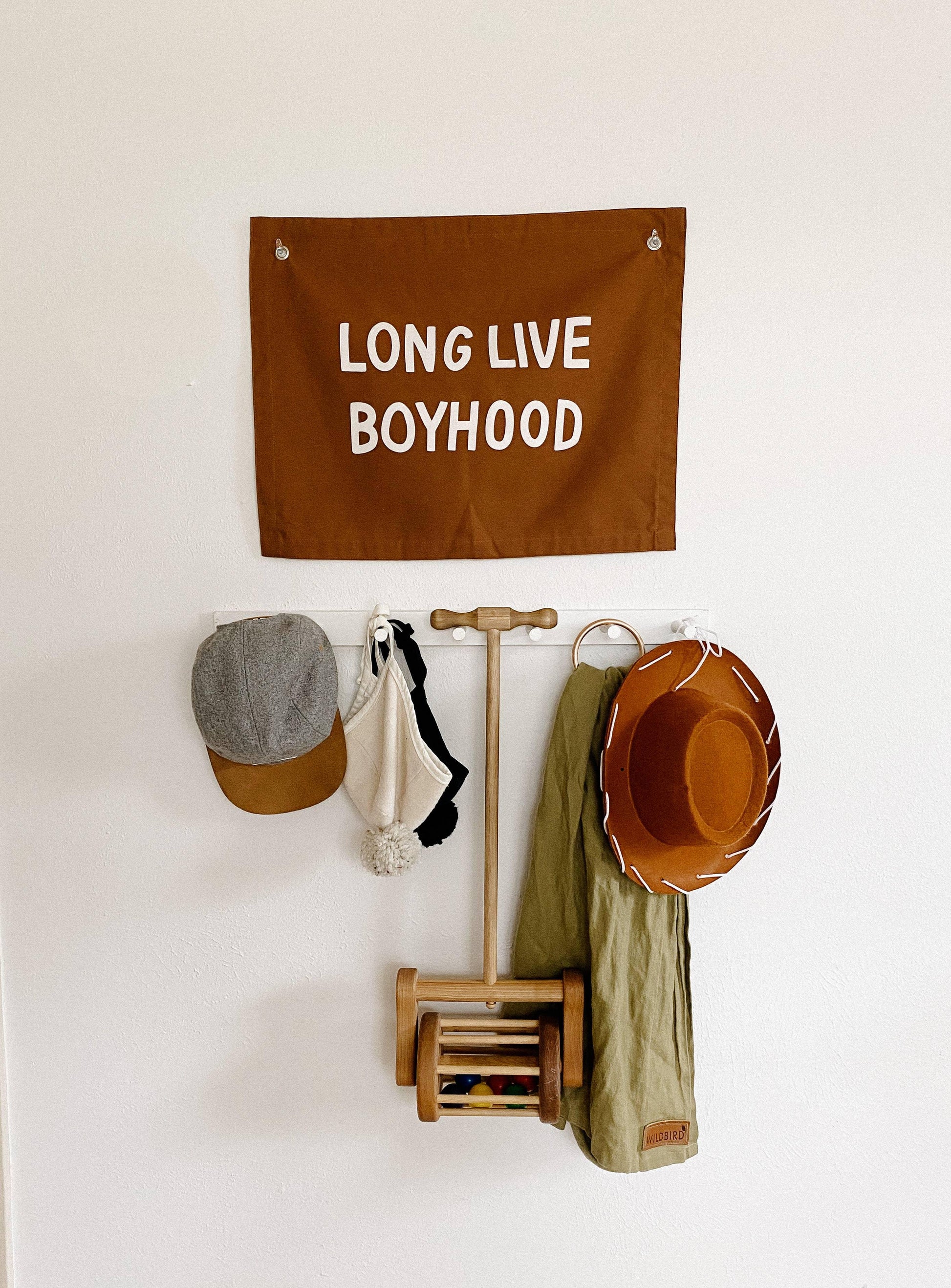 Brown 'Long Live Boyhood' flag, kids hats, scarf, and wooden toy on wall. Come see us at Awaken, Franklin, Tennessee.