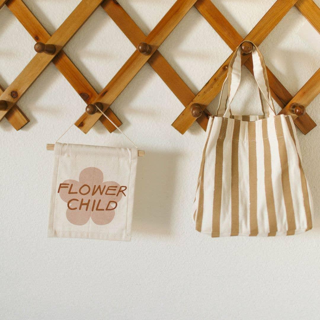 Canvas wall hanging with 'Flower Child' and a striped tote bag on wooden rack, Come see us at Awaken, Franklin, Tennessee